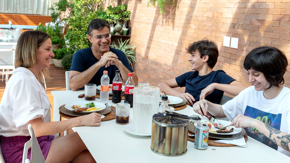Descrição de imagem: fotografia colorida de momento familiar. Rodrigo encabeça a mesa de almoço, Luciana, João Pedro e Isa dividem a mesa, em momento descontraído, todos sorriem. Fim da descrição.