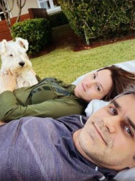 Descrição de imagem: fotografia colorida de momento familiar. Rodrigo e Luciana estão deitados em grama. Rodrigo em primeiro plano, tirando a selfie. Ao fundo, Luciana sorri. Ela está ao lado de Tintim, um cachorro pequeno peludo e branco. Fim da descrição.