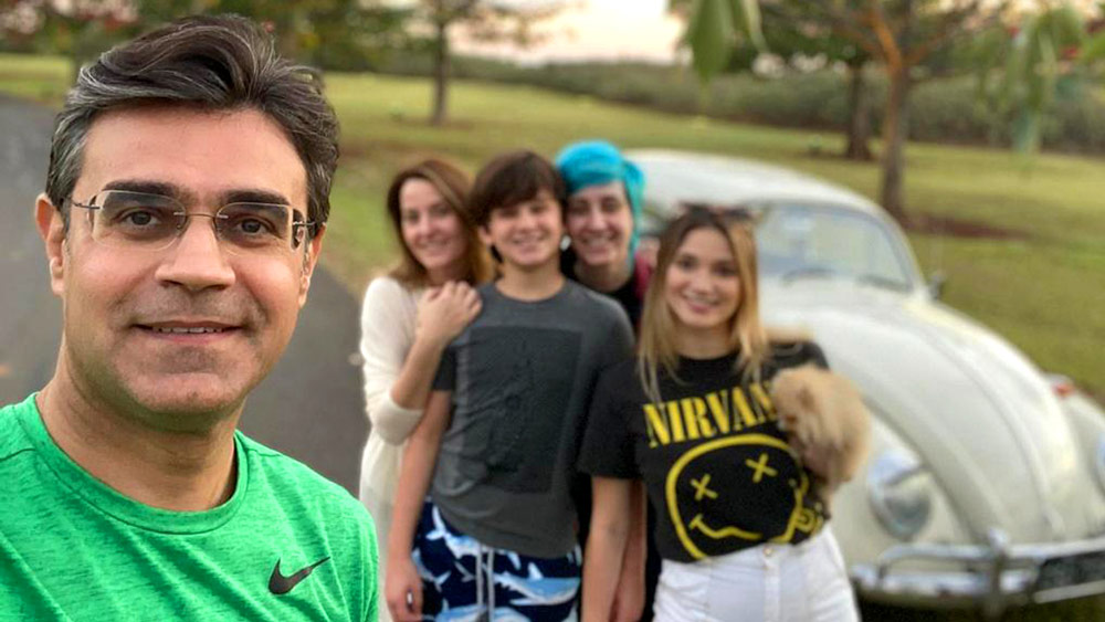 Descrição de imagem: fotografia colorida de momento familiar. Selfie tirada por Rodrigo, que está mais à frente da foto. Ao fundo, abraçados, Luciana, João Pedro, Isa e Valentina. Fim da descrição.