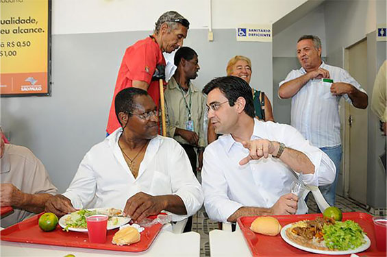 Descrição de imagem: fotografia colorida em ambiente do Bom Prato. Rodrigo está ao lado de homem com pele preta. Ambos estão sentados, com pratos de comida apoiados em mesa. Fim da descrição.